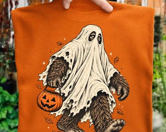 May include: An orange t-shirt featuring a graphic illustration of a brown, furry creature wearing a white ghost sheet. The creature is holding an orange jack-o'-lantern pumpkin bucket, surrounded by small falling autumn leaves and star shapes.
