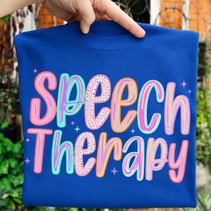 May include: A royal blue t-shirt featuring the text "Speech Therapy" in a multi-coloured, hand-lettered style font. The letters are outlined in pink, orange, and teal, with some filled with black polka dots. Small four-pointed stars surround the text.