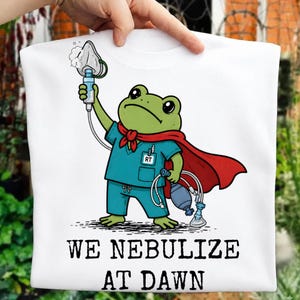 May include: A white t-shirt featuring a green frog dressed in teal medical scrubs and a red cape. The frog holds an oxygen mask and a manual resuscitator. The text "WE NEBULIZE AT DAWN" is printed in black below the illustration of the frog.
