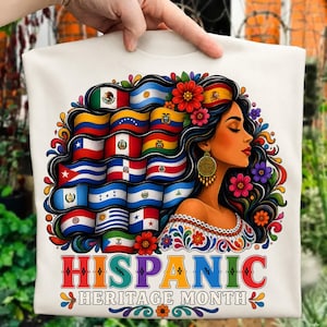 May include: A white t-shirt featuring a graphic of a person with long dark hair styled into various national flags, adorned with colorful flowers and gold earrings. The text HISPANIC HERITAGE MONTH is printed in large, multi-colored letters at the bottom.