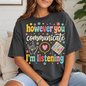 Op de afbeelding: Een antracietgrijs t-shirt met korte mouwen en de kleurrijke tekst "however you communicate I'm listening", een hand die het gebarentaalteken voor "ik hou van jou" maakt, een roze hart in een tekstballon en een communicatiebord.
