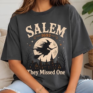 Könnte beinhalten: Ein anthrazitfarbenes T-Shirt mit einem Grafikdruck einer Hexe, die auf einem Besen vor einem großen Vollmond fliegt. Der Text lautet SALEM 1692 und They Missed One, mit einer Silhouette einer Stadt mit Kirchtürmen und kahlen Bäumen.