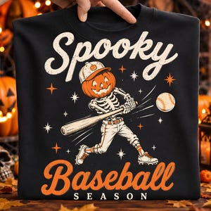 May include: A black crewneck t-shirt featuring a graphic of a skeleton with a pumpkin head wearing a baseball cap and uniform, swinging a wooden bat at a baseball. The text reads Spooky Baseball Season in orange and cream lettering.