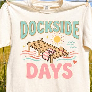 May include: Cream-colored t-shirt with the words "DOCKSIDE DAYS" in a retro font. The design includes a pier, sun, waves, and a beach hat. The shirt is hanging on a clothesline.