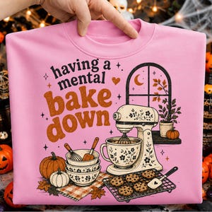 May include: A light pink sweatshirt featuring the text having a mental bake down in black and orange. The graphic includes a stand mixer, stacked bowls, pumpkins, a cooling rack with chocolate chip cookies, and a window with autumn leaves.