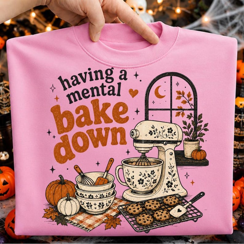 May include: A light pink sweatshirt featuring the text having a mental bake down in black and orange. The graphic includes a stand mixer, stacked bowls, pumpkins, a cooling rack with chocolate chip cookies, and a window with autumn leaves.