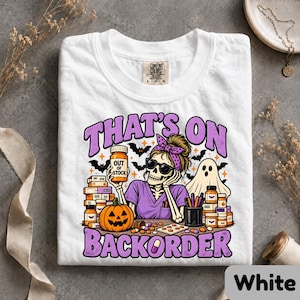 May include: A white t-shirt featuring a graphic of a skeleton wearing a purple scrub top, a headband, and sunglasses. The skeleton holds a pill bottle labeled OUT OF STOCK. The text THAT'S ON BACKORDER surrounds the illustration with pumpkins, ghosts, and medicine bottles.