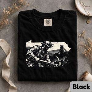 May include: A black t-shirt featuring a white graphic of a miner wearing a hard hat and using a pickaxe, set against the silhouette of the state of Pennsylvania. The graphic includes a mine headframe in the background. The shirt has a Comfort Colors label.