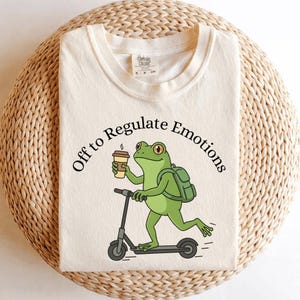 Puede incluir: Camiseta color crema con una rana verde en un patinete, sosteniendo una taza de café. La rana lleva una mochila, y el texto "Off to Regulate Emotions" está encima. La camiseta tiene cuello redondo y mangas cortas.