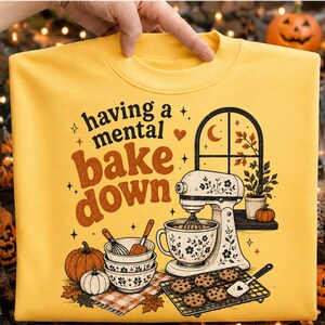 May include: A yellow sweatshirt featuring a graphic print with the text "having a mental bake down," a stand mixer, stacked bowls, a whisk, a spatula, cookies on a cooling rack, pumpkins, and a window view with autumn leaves and a crescent moon.