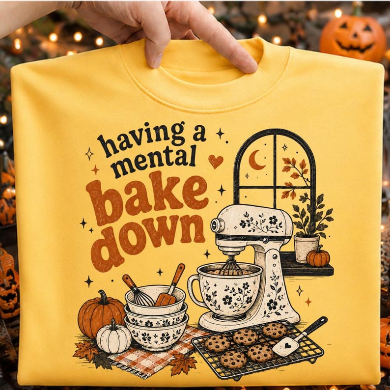 May include: A yellow sweatshirt featuring a graphic print with the text "having a mental bake down," a stand mixer, stacked bowls, a whisk, a spatula, cookies on a cooling rack, pumpkins, and a window view with autumn leaves and a crescent moon.