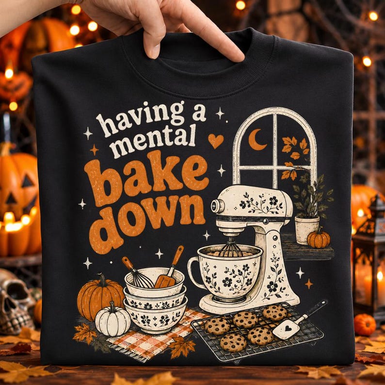 May include: A black sweatshirt featuring a graphic print with the text "having a mental bake down". The design includes a stand mixer, mixing bowls, pumpkins, a cooling rack with cookies, and a window scene with a crescent moon and autumn leaves.