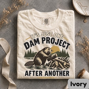 May include: An ivory t-shirt featuring a graphic of a beaver building a dam with a forest and sun in the background. The text reads IT'S JUST ONE DAM PROJECT AFTER ANOTHER. The shirt has a crew neck and a Comfort Colors label on the inside collar.