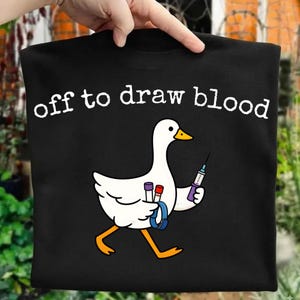 Puede incluir: Una camiseta negra con una oca blanca de dibujos animados con pico y patas naranjas. La oca sostiene una jeringuilla y dos viales médicos, con un torniquete azul alrededor del ala. El texto "off to draw blood" está impreso en blanco arriba.