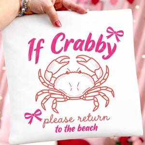 以下が含まれることがあります： ピンク色のカニのイラストと「If Crabby please return to the beach」の文字が入った白い正方形のクッション。カニのデザインには、ハサミと脚のあるカニと2つのリボンが含まれています。テキストは手書き風です。