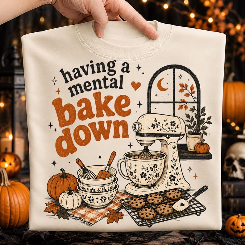 May include: A cream-coloured t-shirt featuring a graphic print with the text "having a mental bake down," a stand mixer, stacked bowls, a cooling rack with cookies, pumpkins, and a window scene with autumn leaves and a crescent moon.