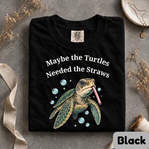 May include: A black crew neck t-shirt featuring a graphic of a sea turtle with a red and white striped straw in its mouth, surrounded by light blue bubbles. The text above the turtle reads, "Maybe the Turtles Needed the Straws" in white.