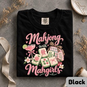 May include: A black t-shirt featuring a graphic with the text "Mahjong with Mahgirls". The design includes a pink cocktail with a lime slice, several mahjong tiles with floral and character motifs, a gift box with a leopard print bow, and cherries.
