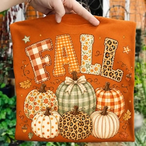 May include: An orange t-shirt featuring the word FALL in large block letters with varied patterns including plaid, floral, and leopard print. Below the text are six pumpkins with different textures, patterns, and a central bow accent.