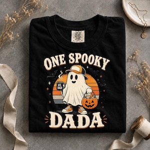 May include: A black t-shirt featuring a graphic of a ghost wearing a baseball cap and trainers, holding a mug labelled #1 DAD and a jack-o'-lantern bucket. The text reads ONE SPOOKY DADA against an orange sunset background with bats and stars.