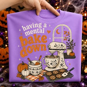 May include: A light purple crewneck sweatshirt featuring a graphic print with the text having a mental bake down. The design includes a stand mixer, stacked mixing bowls, a whisk, a spatula, a cooling rack with cookies, and pumpkins.