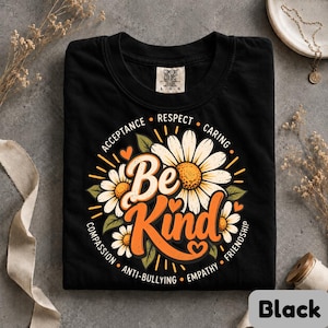 May include: A black t-shirt featuring a graphic of white daisies with yellow centers and green leaves. The text "Be Kind" is printed in orange, surrounded by the words "Acceptance," "Respect," "Caring," "Friendship," "Empathy," "Anti-bullying," and "Compassion."