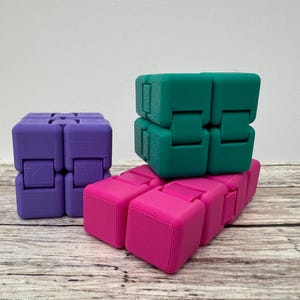 May include: Three infinity cubes in purple, teal, and bright pink. Each cube is composed of eight smaller connected blocks that can be folded and manipulated. The purple cube stands upright on the left, while the teal and pink cubes are stacked.