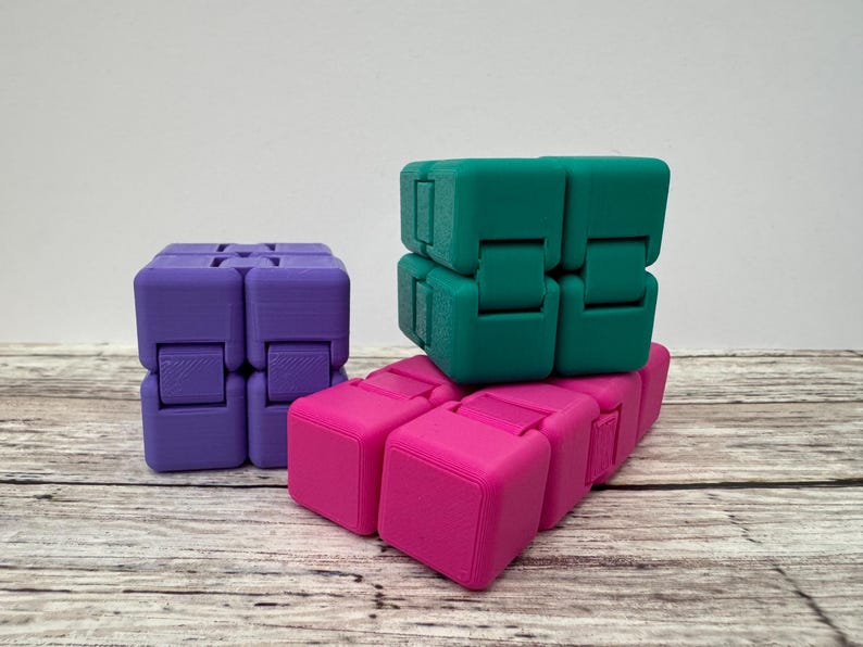 May include: Three infinity cubes in purple, teal, and bright pink. Each cube is composed of eight smaller connected blocks that can be folded and manipulated. The purple cube stands upright on the left, while the teal and pink cubes are stacked.