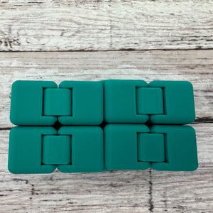 May include: A teal-colored infinity cube fidget toy composed of eight interconnected square blocks. The device features a smooth, matte finish and is designed with multiple hinges that allow the blocks to fold and rotate into various shapes.