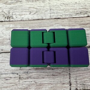 May include: A rectangular infinity cube fidget toy composed of eight interconnected blocks. The top row consists of four solid green blocks, while the bottom row features four solid purple blocks. The blocks are linked by hinges that allow for folding.