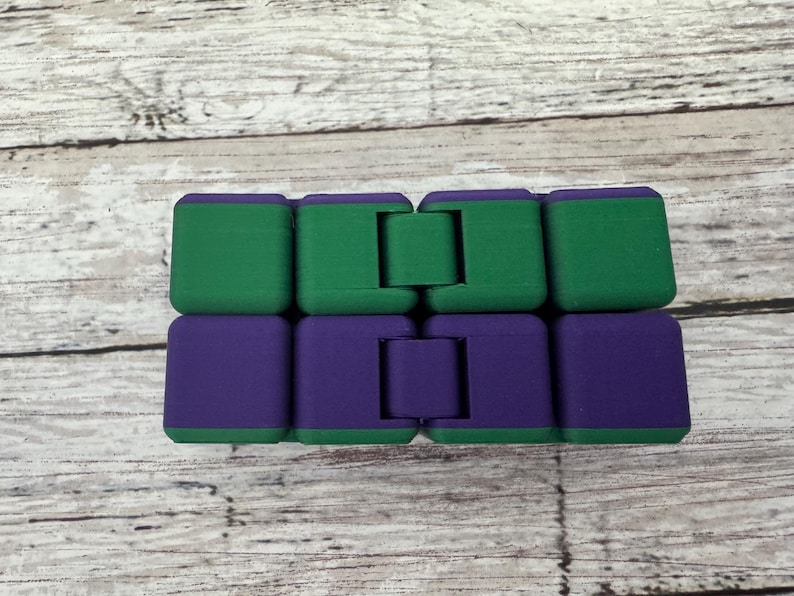 May include: A rectangular infinity cube fidget toy composed of eight interconnected blocks. The top row consists of four solid green blocks, while the bottom row features four solid purple blocks. The blocks are linked by hinges that allow for folding.
