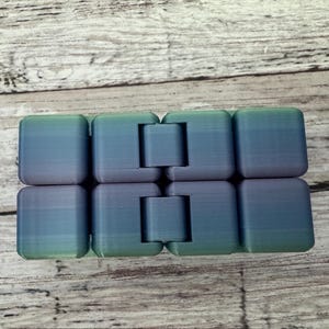 May include: An infinity cube fidget toy featuring a gradient color scheme that transitions from green to blue and purple. The object is composed of eight interconnected plastic blocks with visible hinges that allow the segments to fold and rotate.