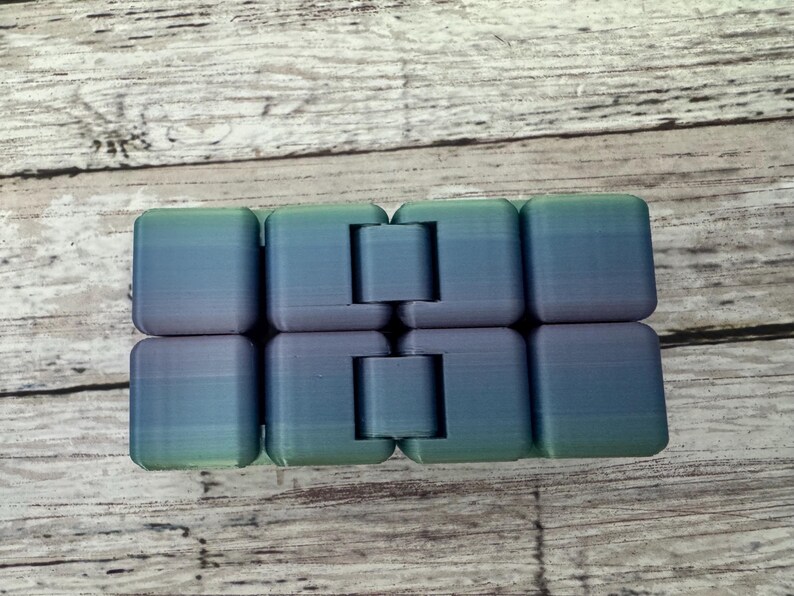 May include: An infinity cube fidget toy featuring a gradient color scheme that transitions from green to blue and purple. The object is composed of eight interconnected plastic blocks with visible hinges that allow the segments to fold and rotate.