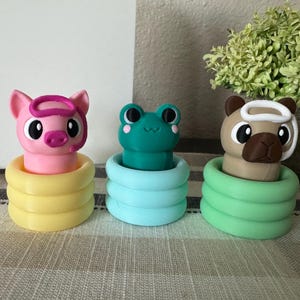 May include: Three colorful animal-themed bottle stoppers. A pink pig with a pink headband sits atop a yellow base. A teal frog sits on a light blue base. A tan llama sits on a green base.