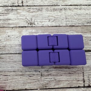 May include: A purple infinity cube fidget toy composed of eight small, interconnected square blocks. The device features a smooth, matte finish and visible hinges that allow the blocks to fold and rotate into various configurations for tactile play.