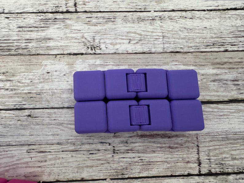 May include: A purple infinity cube fidget toy composed of eight small, interconnected square blocks. The device features a smooth, matte finish and visible hinges that allow the blocks to fold and rotate into various configurations for tactile play.