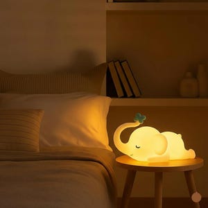 May include: A soft yellow elephant-shaped night light with a small blue flower on its trunk rests on a small wooden table. The lamp is glowing, casting a warm light in a bedroom setting. The elephant lamp is a children's room decor item.