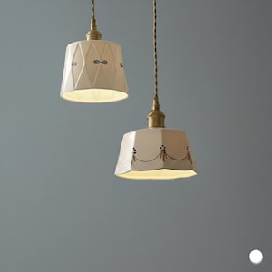 May include: Two cream-colored pendant lights with brass hardware and decorative patterns. One shade features a diamond pattern with blue accents, the other has a floral design. The lights hang from twisted brown cords.