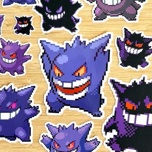 May include: A collection of pixel art stickers featuring the character Gengar in various poses and colour variations, including shades of purple, black, and red. Each sticker displays the character with a wide, toothy grin and pointed ears.