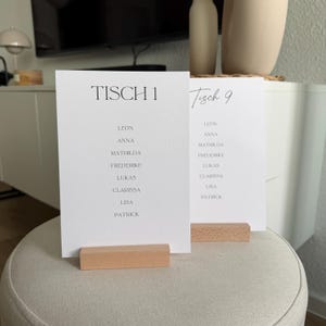 K&ouml;nnte beinhalten: Zwei wei&szlig;e, rechteckige Papierkarten mit schwarzem Text, die jeweils von einem hellen Holzblockhalter aufrecht gehalten werden. Die vordere Karte zeigt den Text TISCH 1 und eine Namensliste, die zweite Karte zeigt Tisch 9 und eine Namensliste.