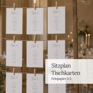 Könnte beinhalten: Ein Sitzplan mit weißen, rechteckigen Karten von Tisch 1 bis 8, die jeweils Namen in schwarzer Schrift auflisten. Die Karten sind mit hölzernen Wäscheklammern an horizontalen Hanfschnüren befestigt, die an einem Holzrahmen angebracht sind.