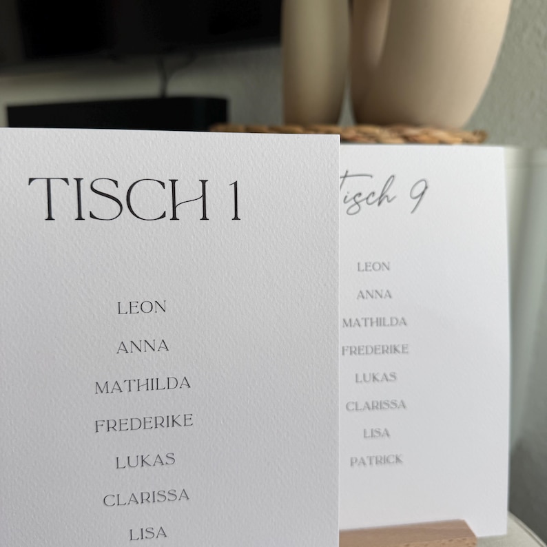 K&ouml;nnte beinhalten: Zwei wei&szlig;e, rechteckige Tischkarten aus strukturiertem Papier. Die vordere Karte zeigt den Text TISCH 1 gefolgt von einer Namensliste: Leon, Anna, Mathilda, Frederike, Lukas, Clarissa und Lisa. Die zweite Karte zeigt die Aufschrift Tisch 9 mit Namen.