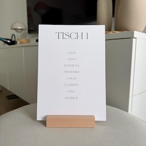 K&ouml;nnte beinhalten: Eine wei&szlig;e rechteckige Karte mit schwarzem Text TISCH 1, gefolgt von einer Namensliste: Leon, Anna, Mathilda, Frederike, Lukas, Clarissa, Lisa und Patrick. Die Karte wird von einem rechteckigen, hellen Holzblock als St&auml;nder aufrecht gehalten.
