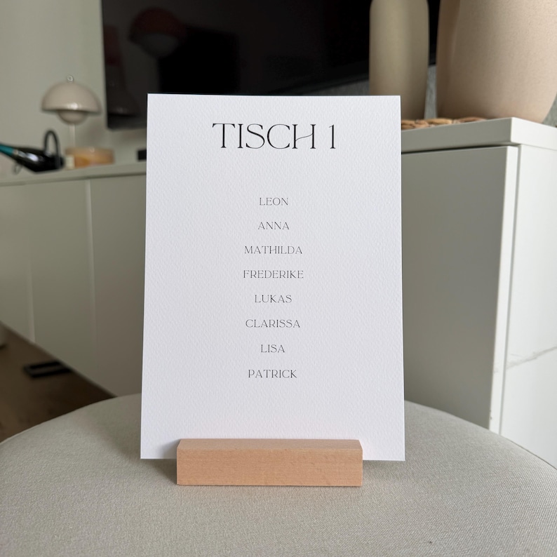 K&ouml;nnte beinhalten: Eine wei&szlig;e rechteckige Karte mit schwarzem Text TISCH 1, gefolgt von einer Namensliste: Leon, Anna, Mathilda, Frederike, Lukas, Clarissa, Lisa und Patrick. Die Karte wird von einem rechteckigen, hellen Holzblock als St&auml;nder aufrecht gehalten.