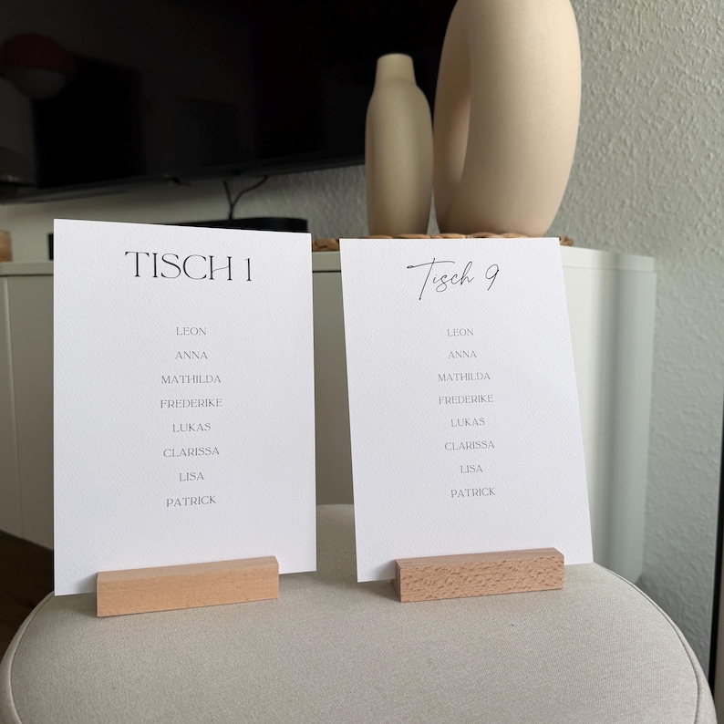 K&ouml;nnte beinhalten: Zwei rechteckige wei&szlig;e Tischkarten aus Papier, die von hellen Holzbl&ouml;cken aufrecht gehalten werden. Die linke Karte zeigt den Text TISCH 1, die rechte Karte den Text Tisch 9. Beide Karten listen die Namen Leon, Anna, Mathilda, Frederike, Lukas, Clarissa, Lisa und Patrick in schwarzer Schrift auf.