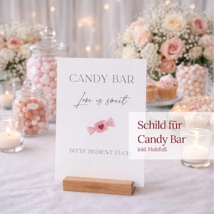 Könnte beinhalten: Ein weißes Schild mit den Worten "CANDY BAR" und "Love is sweet" in eleganter Schrift, mit einer rosa Süßigkeiten-Grafik. Das Schild wird von einem Holzsockel gehalten, ideal für eine Candy Bar-Anzeige.