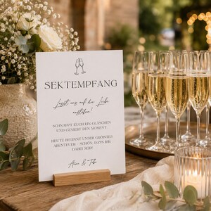以下が含まれることがあります： SEKTEMPFANG、Lasst uns auf die Liebe ansto&szlig;en!、SCHNAPPT EUCH EIN GL&Auml;SCHEN UND GENIEBT DEN MOMENT、HEUTE BEGINNT UNSER GR&Ouml;ISTES ABENTEUER、Anna & Tobiと黒い文字で書かれた白い長方形の看板。明るい木製のブロックスタンドに立てられている。