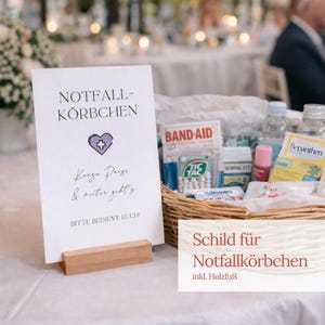Könnte beinhalten: Ein Schild mit den Worten "NOTFALL-KORBCHEN" und einem lila Herzsymbol, das auf einem Holzständer steht. Ein Weidenkorb mit Erste-Hilfe-Materialien, darunter Pflaster und Tic Tacs, ist im Hintergrund zu sehen.