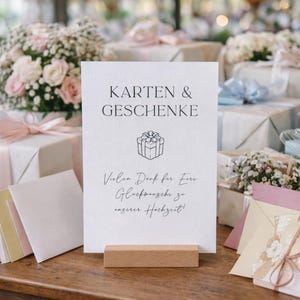Könnte beinhalten: Ein weißes Schild mit den Worten "Karten & Geschenke" und einer Geschenkbox-Illustration. Das Schild wird von einem Holzständer gehalten. Der Hintergrund zeigt verpackte Geschenke und Blumengestecke.