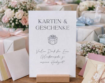 Könnte beinhalten: Ein weißes Schild mit den Worten "Karten & Geschenke" und einer Geschenkbox-Illustration. Das Schild wird von einem Holzständer gehalten. Der Hintergrund zeigt verpackte Geschenke und Blumengestecke.